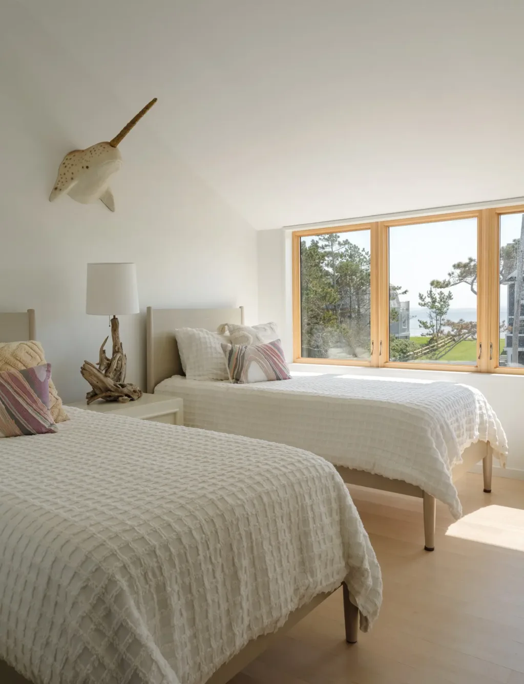 Bright bedroom with two twin beds, textured white bedding, colorful pillows, a window with a view, and a decorative fish mounted on the white wall.