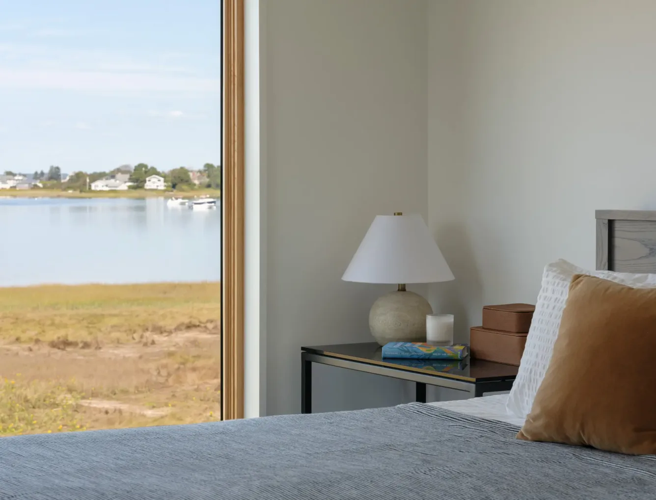 A cozy bedroom with a neatly made bed, a side table holding a lamp and books, and a large window overlooking a calm lake and houses in the distance.