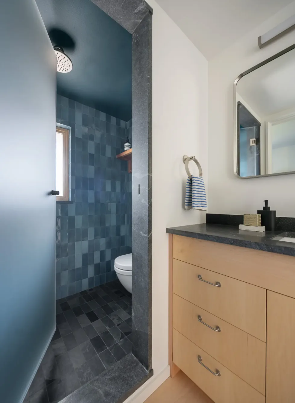 Modern bathroom with a blue-tiled walk-in shower, wooden vanity with black countertop, rectangular mirror, and striped hand towel on a ring holder.