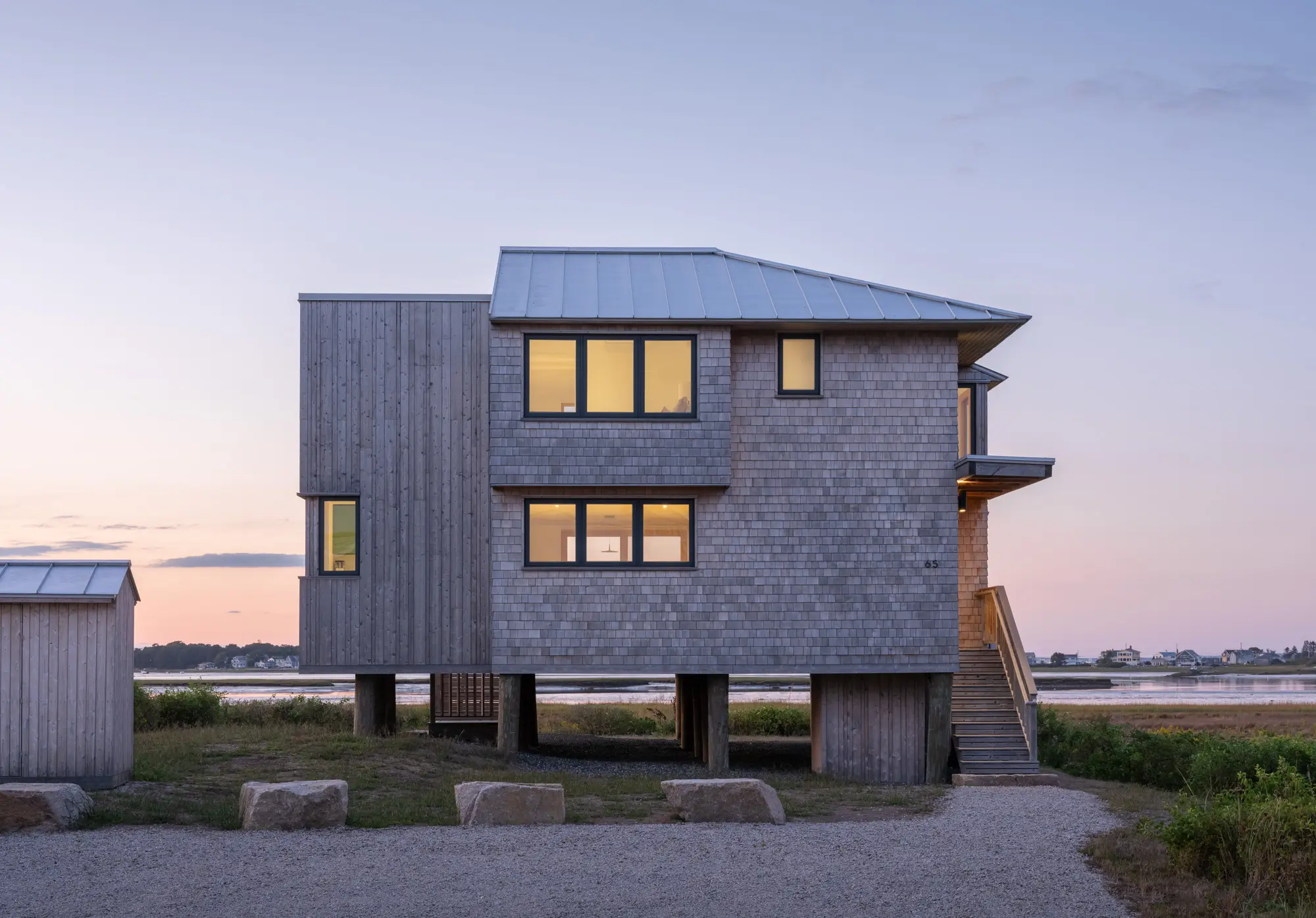 Modern wooden house on stilts with large windows, situated near water at sunset with a gravel path and grassy surroundings.