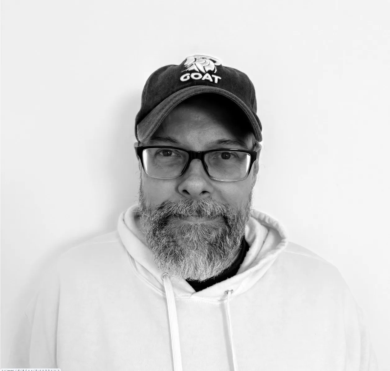 A bearded man wearing glasses, a hoodie, and a baseball cap with the word GOAT stands against a plain white background.