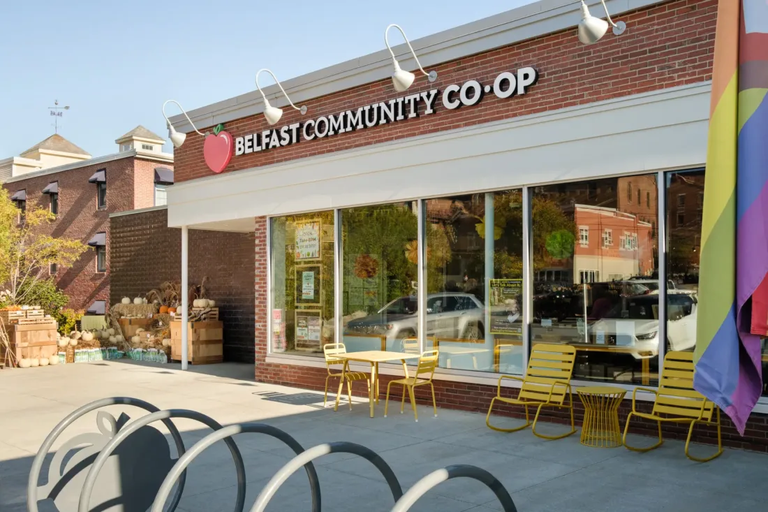 Exterior of the Belfast Community Co-op with yellow chairs and tables outside, large windows, and a rainbow flag visible on the right.