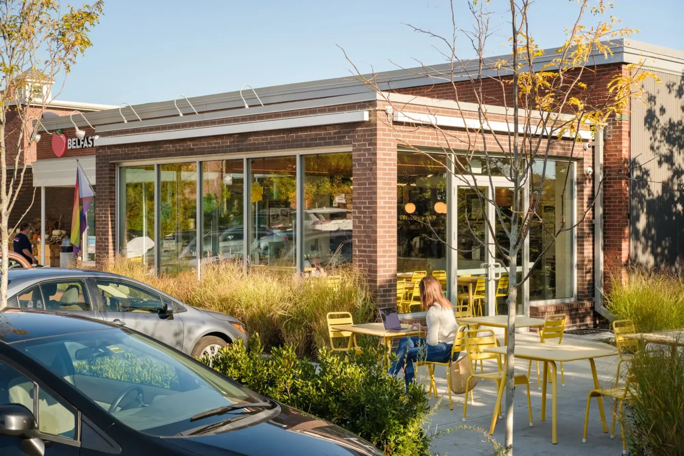 A woman sits at an outdoor table with a laptop at a brick café with large windows and yellow chairs; cars are parked nearby and trees surround the area.
