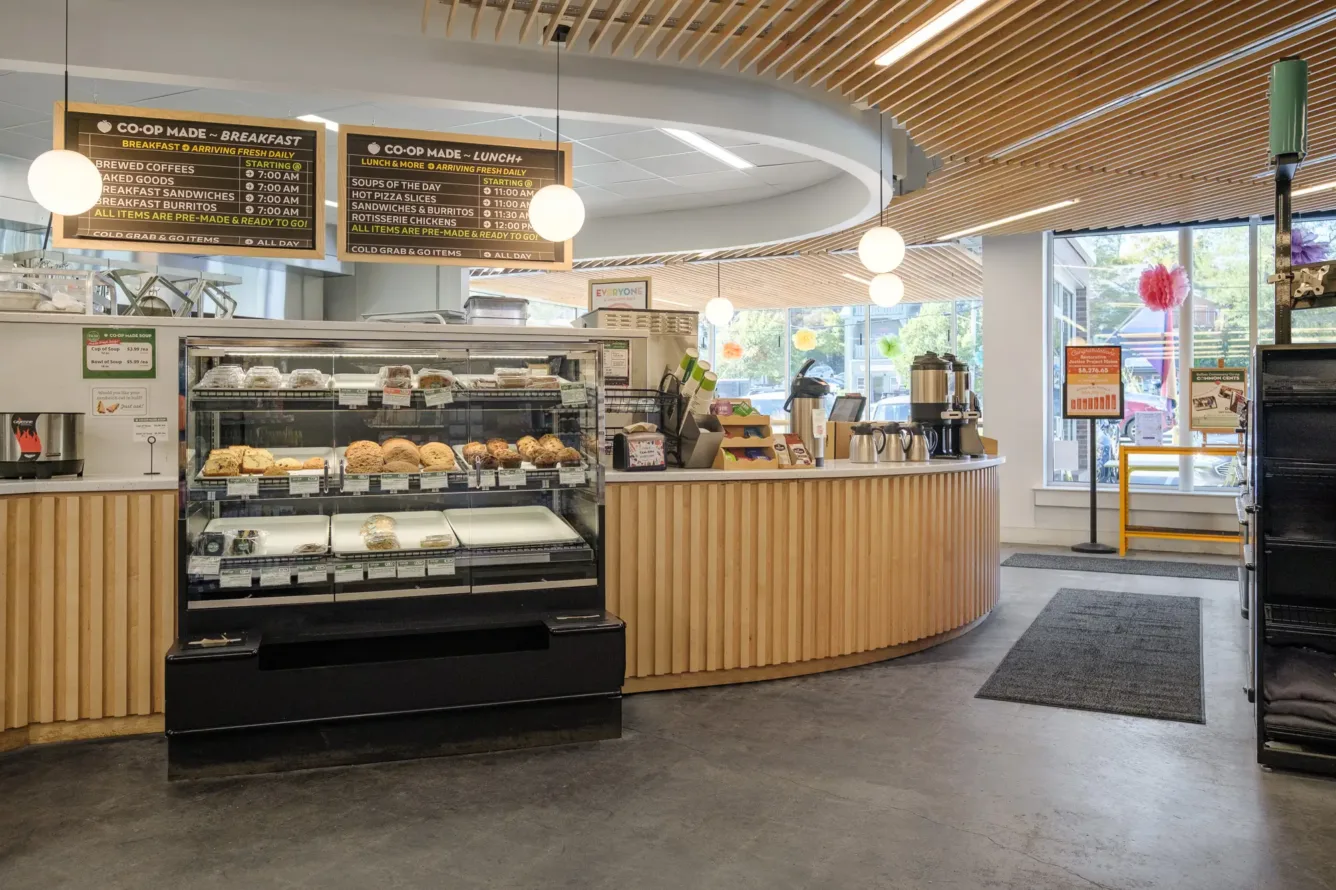 A bakery café with a pastry display case, a curved wooden counter, hanging menus, coffee dispensers, and a modern, well-lit interior.