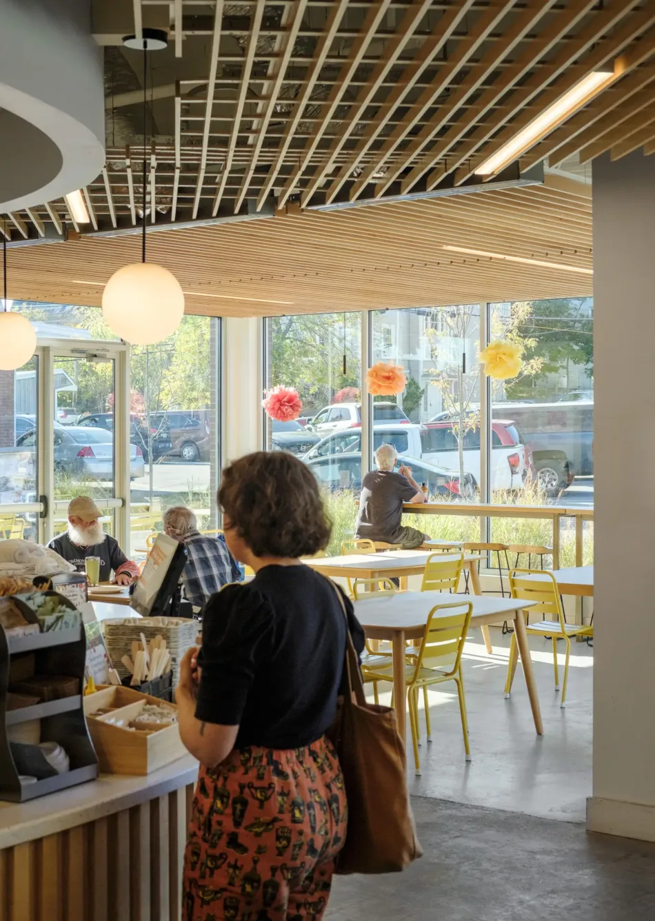 A woman stands at a café counter while people sit at tables near large windows with colorful decorations and sunlight streaming in.