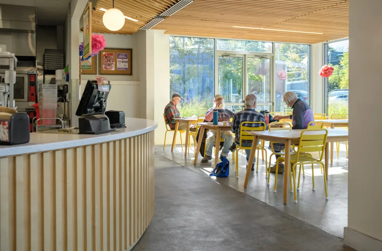 Four older adults sit at a table with yellow chairs in a bright, modern café with large windows and wooden ceiling slats. Sunlight streams in through the glass doors.