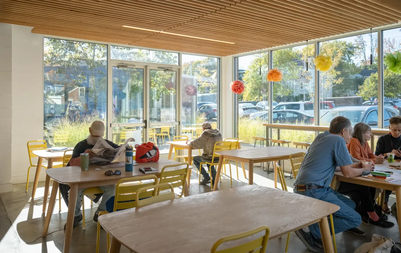 A bright café with yellow chairs, wooden tables, and large windows. Several people are eating, reading, or talking. Pom-pom decorations hang in the window. Sunlight fills the room.