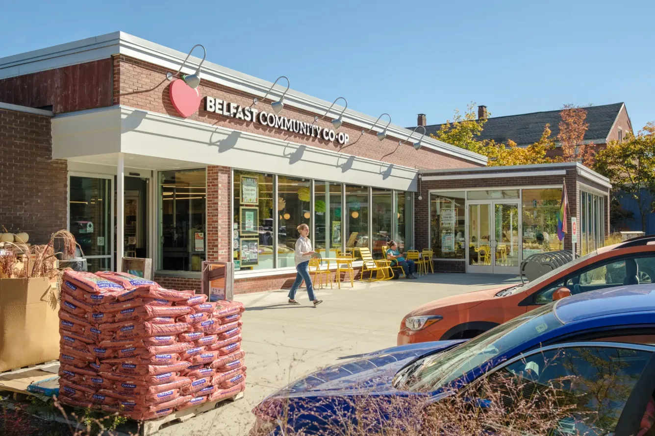 A person walks in front of Belfast Community Co-op, with stacked bags, parked cars, and yellow outdoor seating visible on a sunny day.