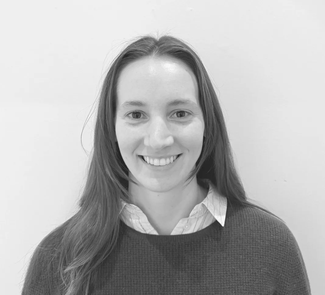 A woman with long, straight hair smiles at the camera. She is wearing a collared shirt under a sweater and is standing against a plain, light-colored background.