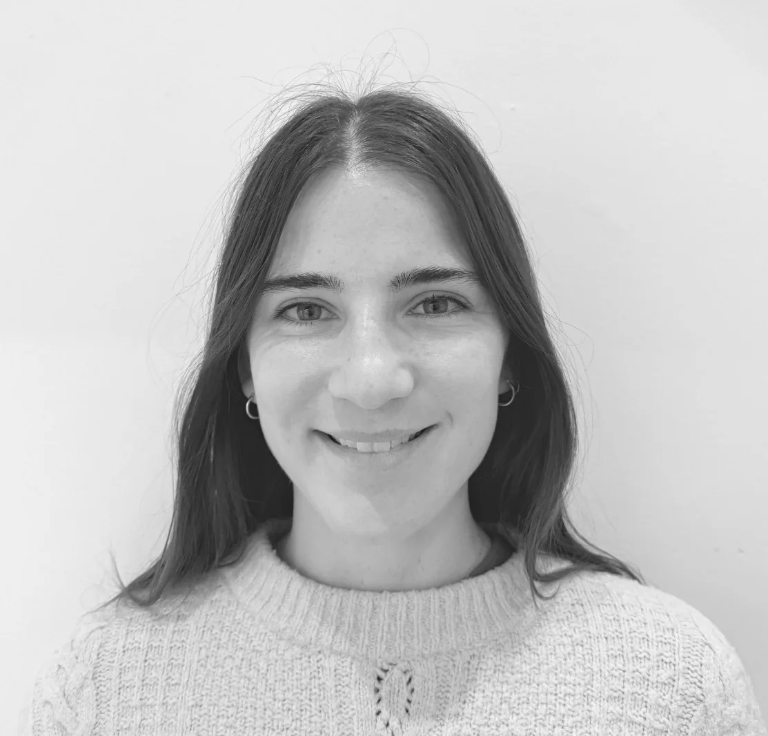 A young woman with straight, dark hair and hoop earrings smiles at the camera. She is wearing a light-colored knit sweater against a plain white background.