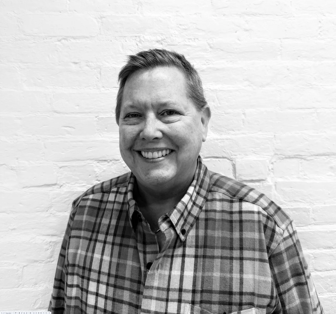 A person with short hair, wearing a plaid shirt, smiling while standing in front of a white brick wall.