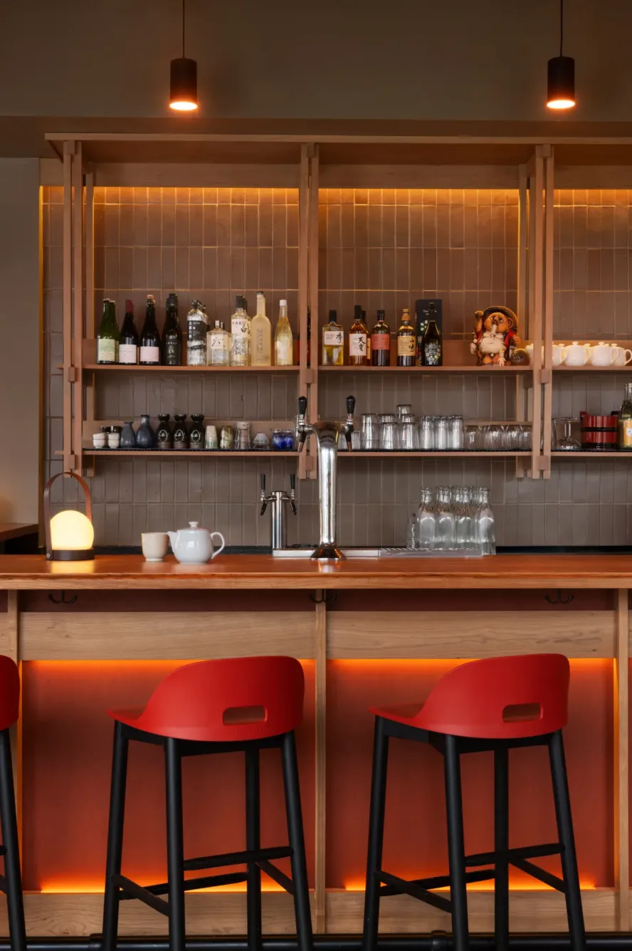 A modern bar with wooden shelves, bottles, glasses, and teapots, warm lighting, and two red bar stools at a wooden counter.