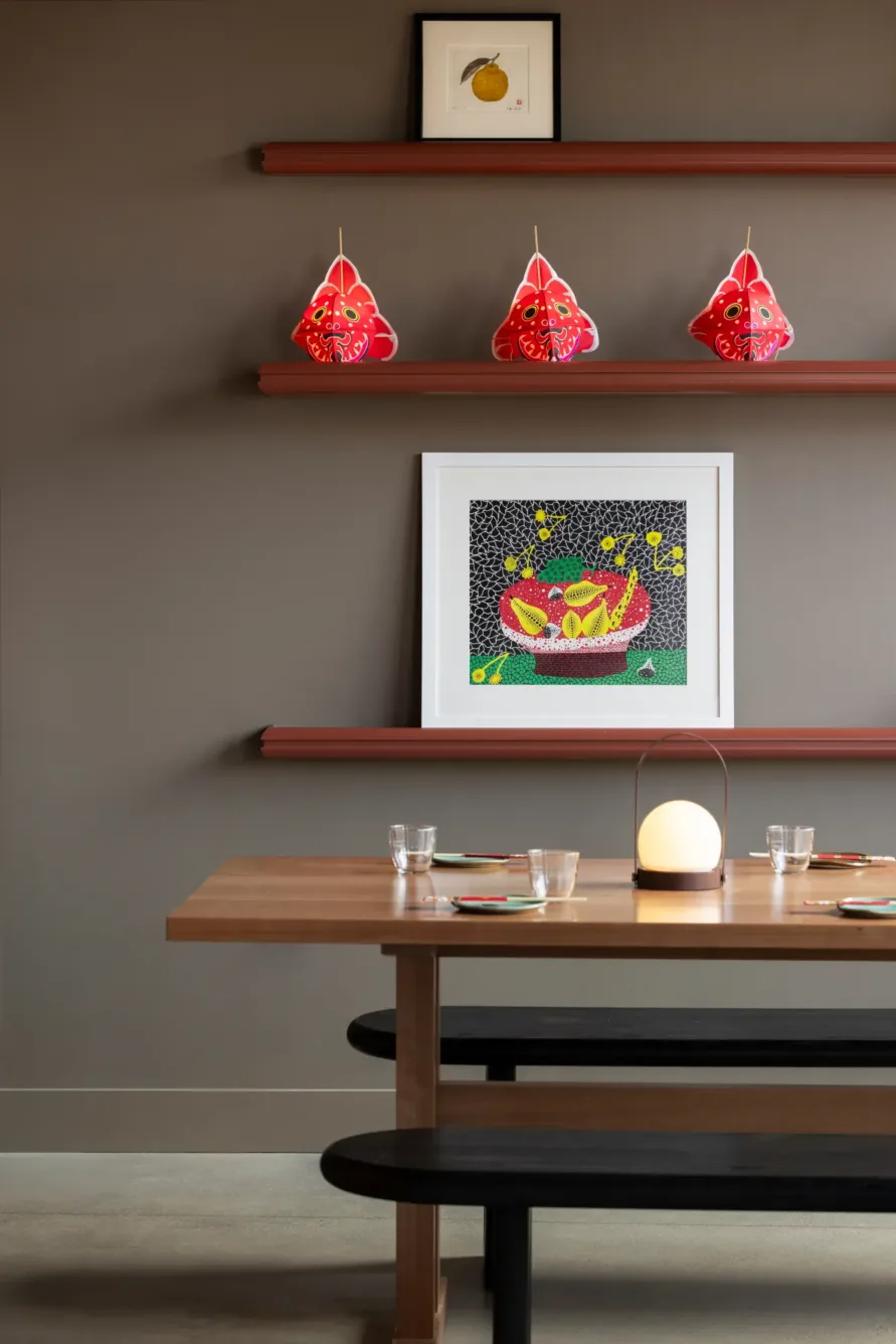 A modern dining area with a wooden table, black bench, glassware, wall art, three red hanging lanterns, and minimalist decor on brown shelves.