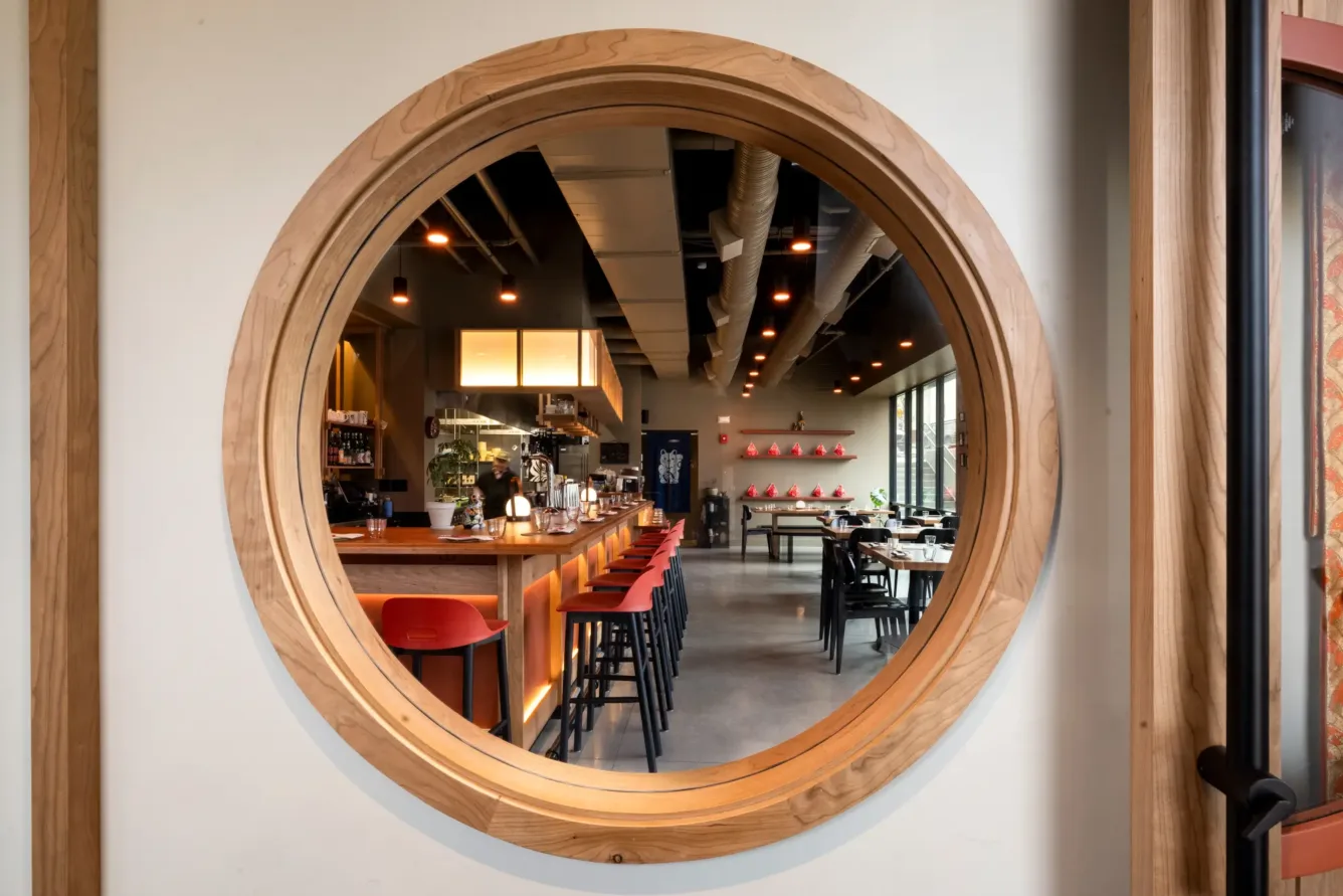View of a modern restaurant interior with a bar, red stools, and dining tables, seen through a large circular wooden window. Warm lighting creates a cozy atmosphere.