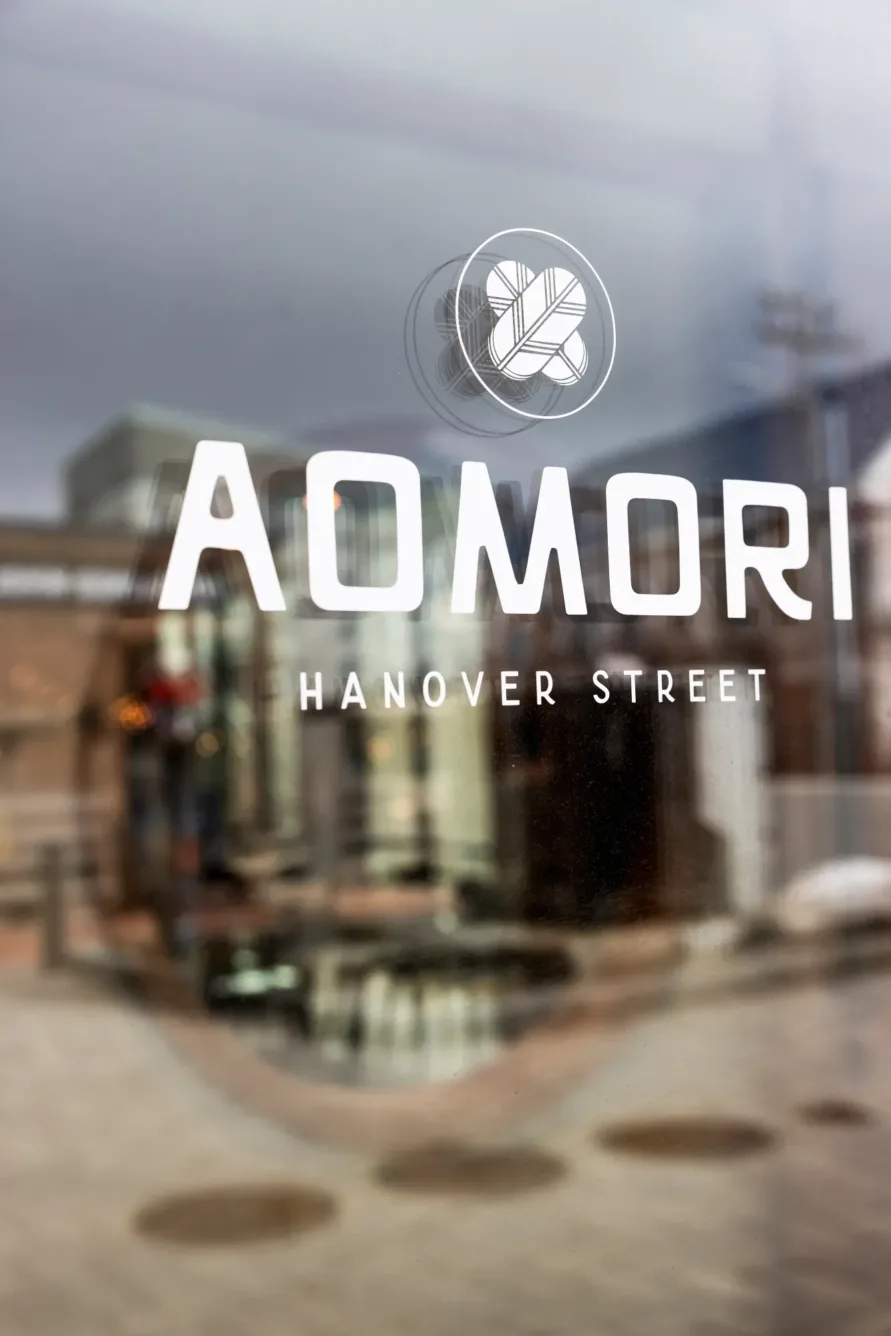 A glass door with the white logo and text AOMORI HANOVER STREET reflecting buildings and a cloudy sky.