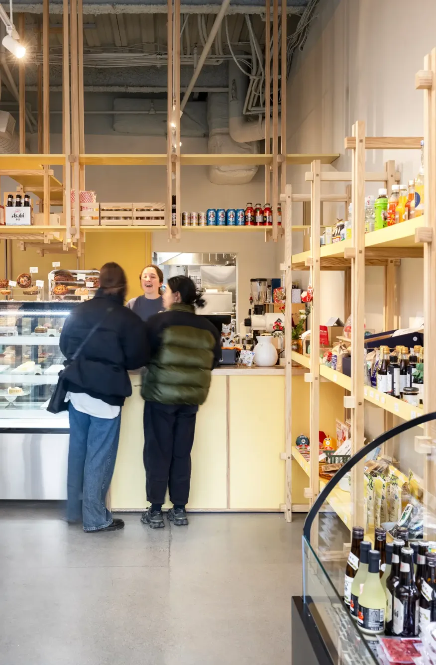 Two people stand at a counter speaking to a barista inside a modern, well-lit café with shelves of food, drinks, and products on display.