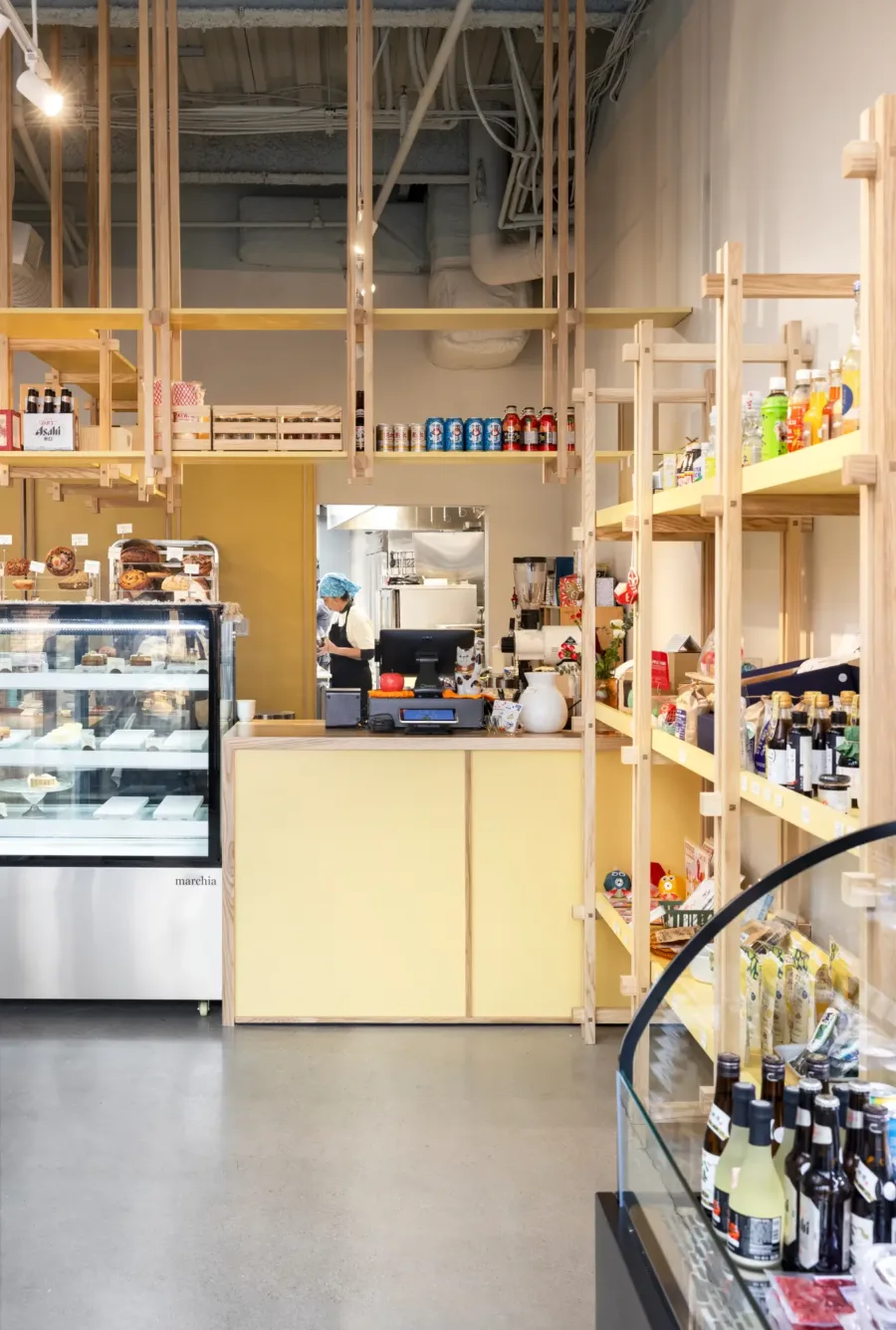 A bright, modern shop interior with wooden shelves displaying snacks and drinks, a glass bakery case, and a person working behind a yellow counter.