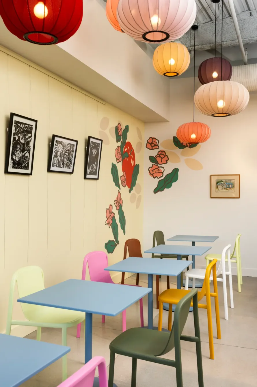 A colorful cafe with pastel chairs, blue tables, hanging lantern lights, wall art of flowers, and framed pictures on a cream wall.