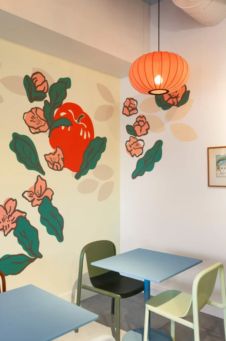 A small cafe corner with pastel blue tables, mismatched chairs, a red pendant lamp, and a wall mural featuring flowers, green leaves, and a large red fruit.