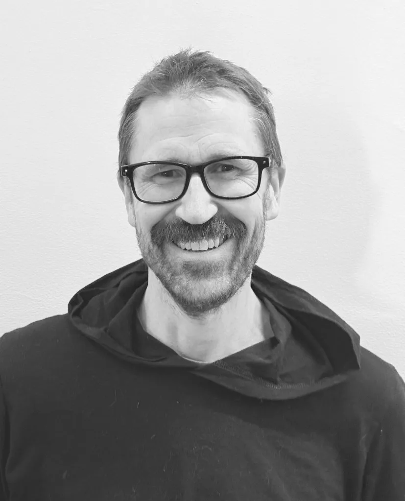 A man with short hair, glasses, and a mustache smiles at the camera. He is wearing a dark hoodie and standing against a plain light-colored background.