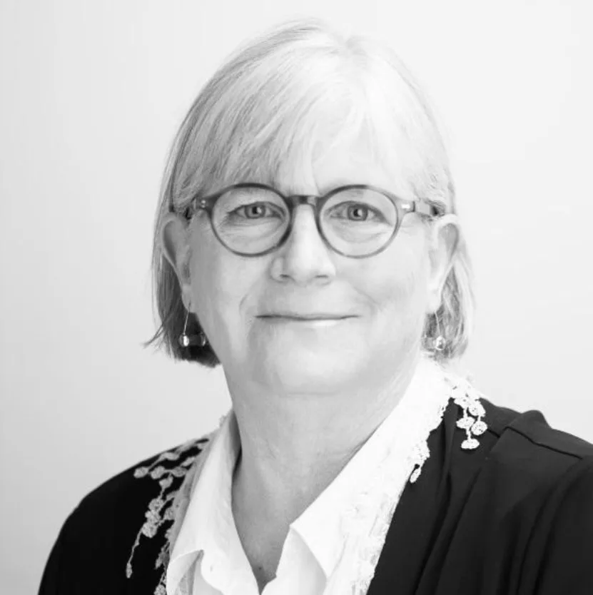 Black and white portrait of an older woman with light hair and glasses, wearing a dark jacket with decorative trim over a light blouse, smiling slightly at the camera.