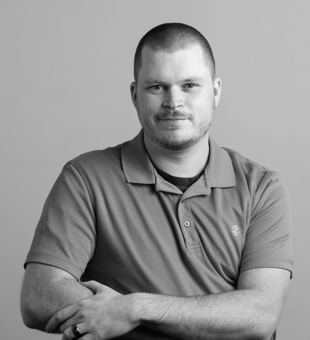 A man with short hair and a trimmed beard, wearing a short-sleeved polo shirt, sits with arms crossed and smiles slightly against a plain background.