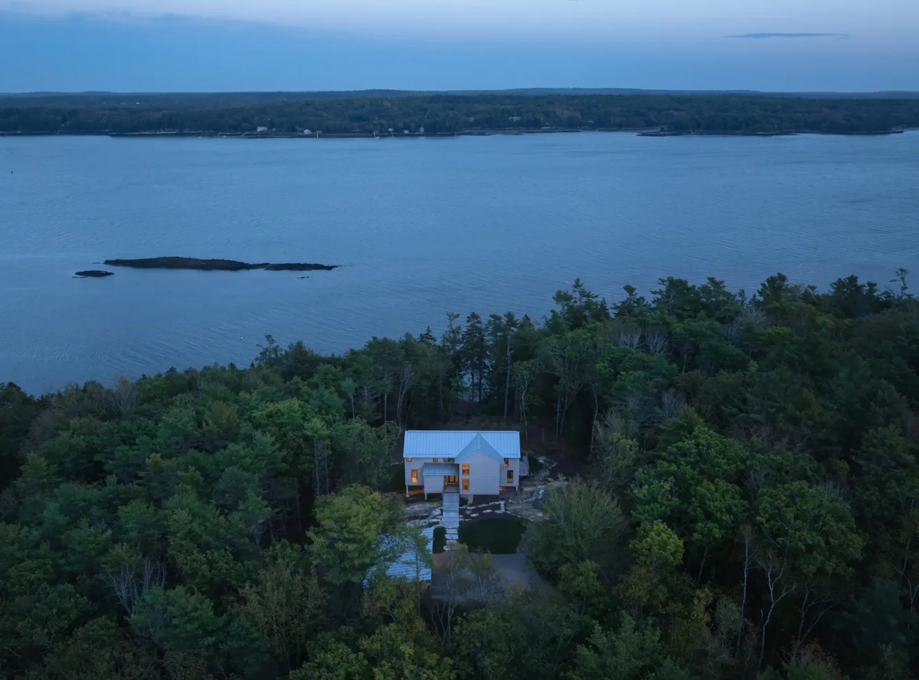 A house with a gray roof sits surrounded by dense trees near the edge of a large, calm body of water at dusk.