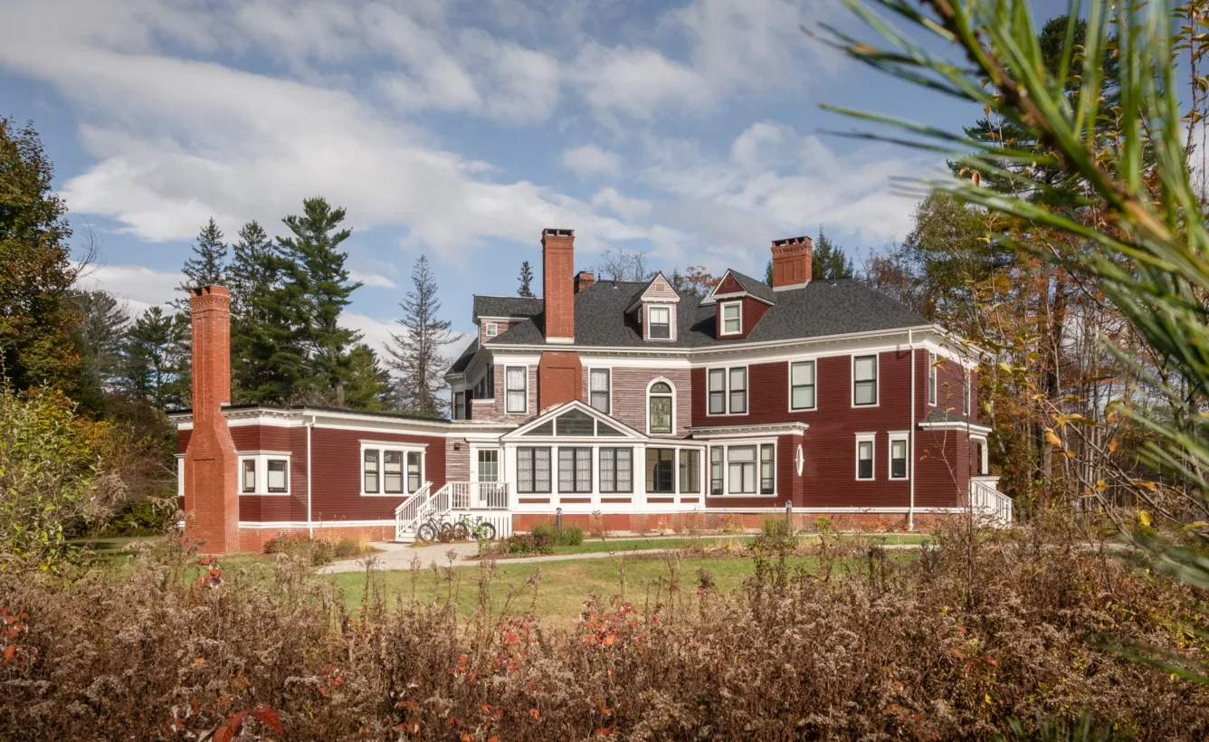 A large, red Victorian-style house with white trim, multiple chimneys, and a glass-enclosed porch, surrounded by trees and shrubs under a partly cloudy sky.