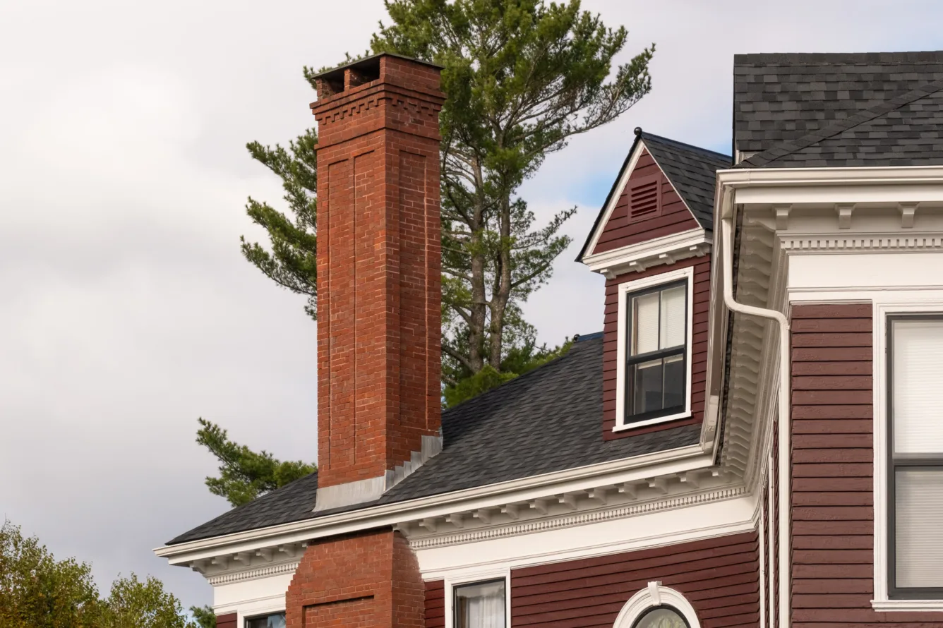 A close-up of a red brick chimney on the roof of a maroon house with white trim, in front of a tall pine tree and cloudy sky.