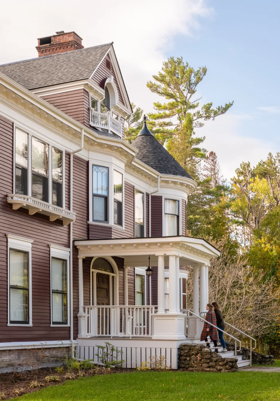 A couple walks up the steps to the porch of a large Victorian-style house with purple siding and white trim, surrounded by trees and greenery.