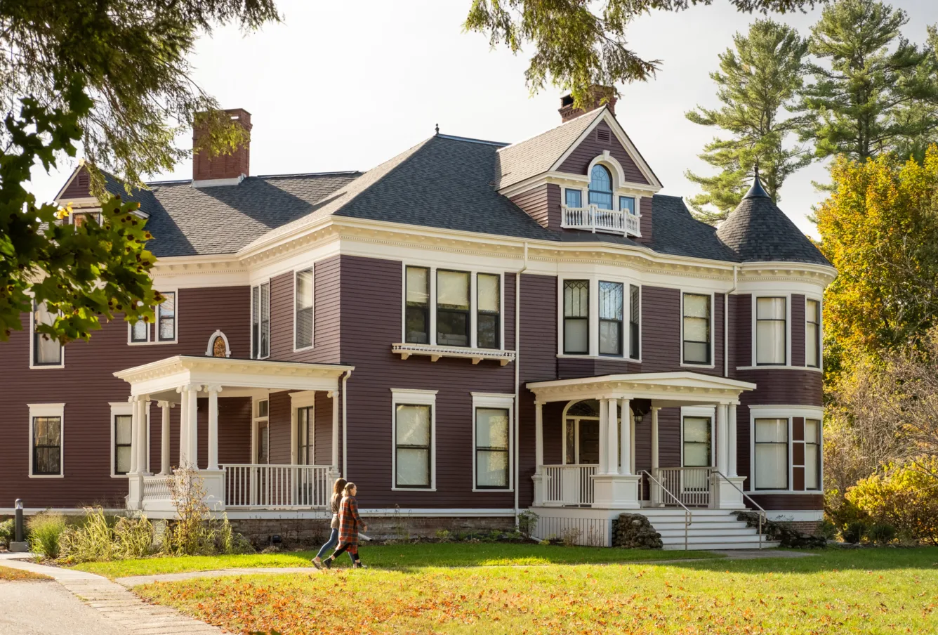 A large, purple Victorian-style house with white trim and a porch; two people walk along the front yard on a sunny day.
