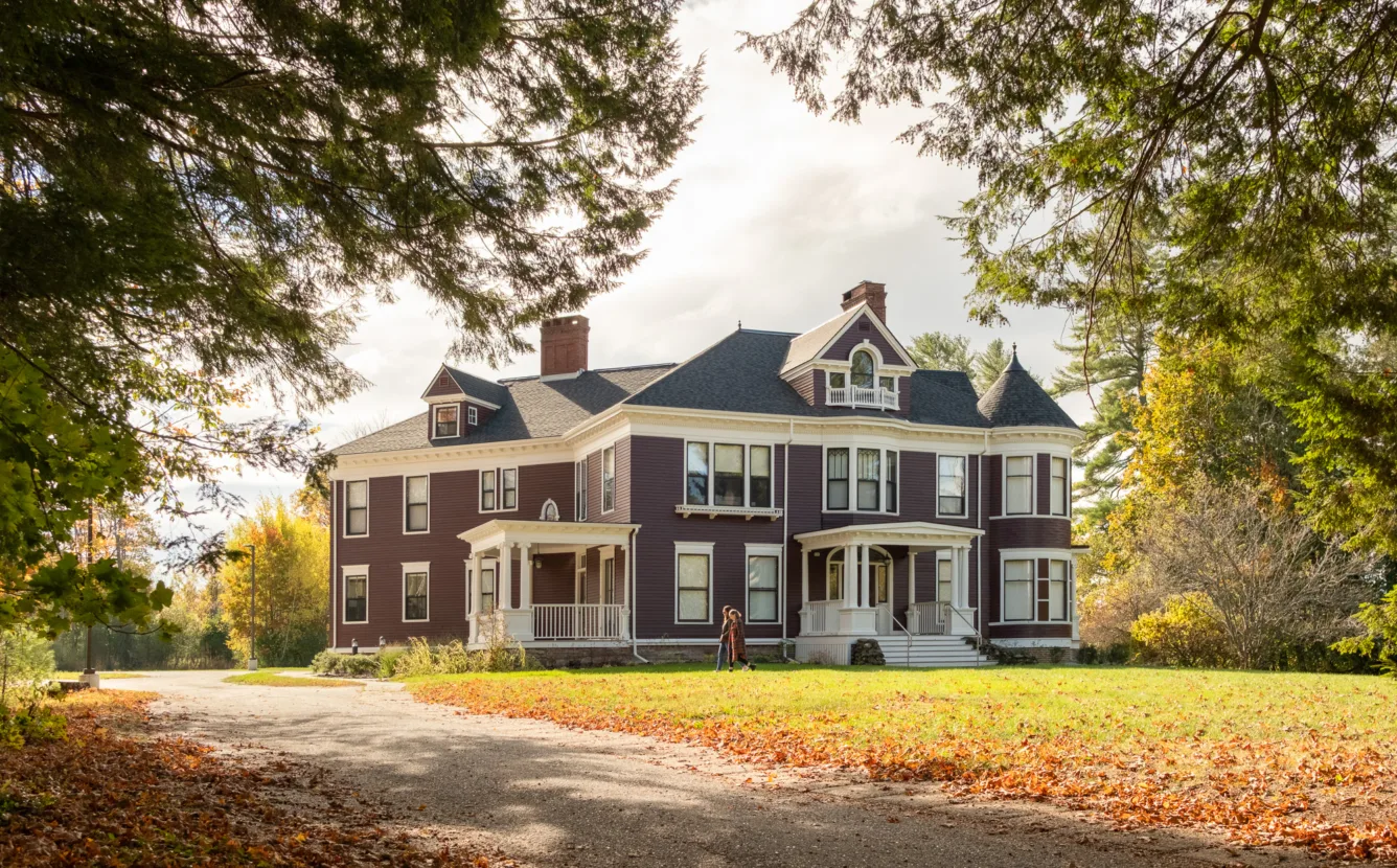 A large, two-story Victorian house with a wraparound porch, set in a grassy yard surrounded by trees and autumn leaves on the ground.