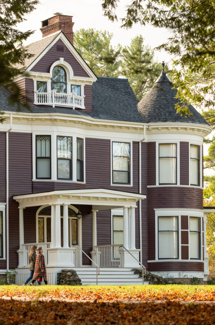 Two people walk past a large, Victorian-style purple house with white trim, rounded turret, and a covered porch, surrounded by trees and fallen autumn leaves.