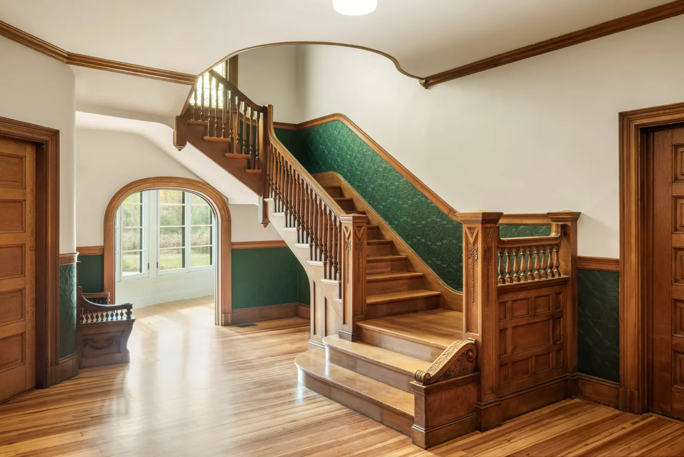 A wooden staircase with ornate railing leads upward in a bright room with green and white walls, wood trim, and polished floors. A large window lets in natural light.
