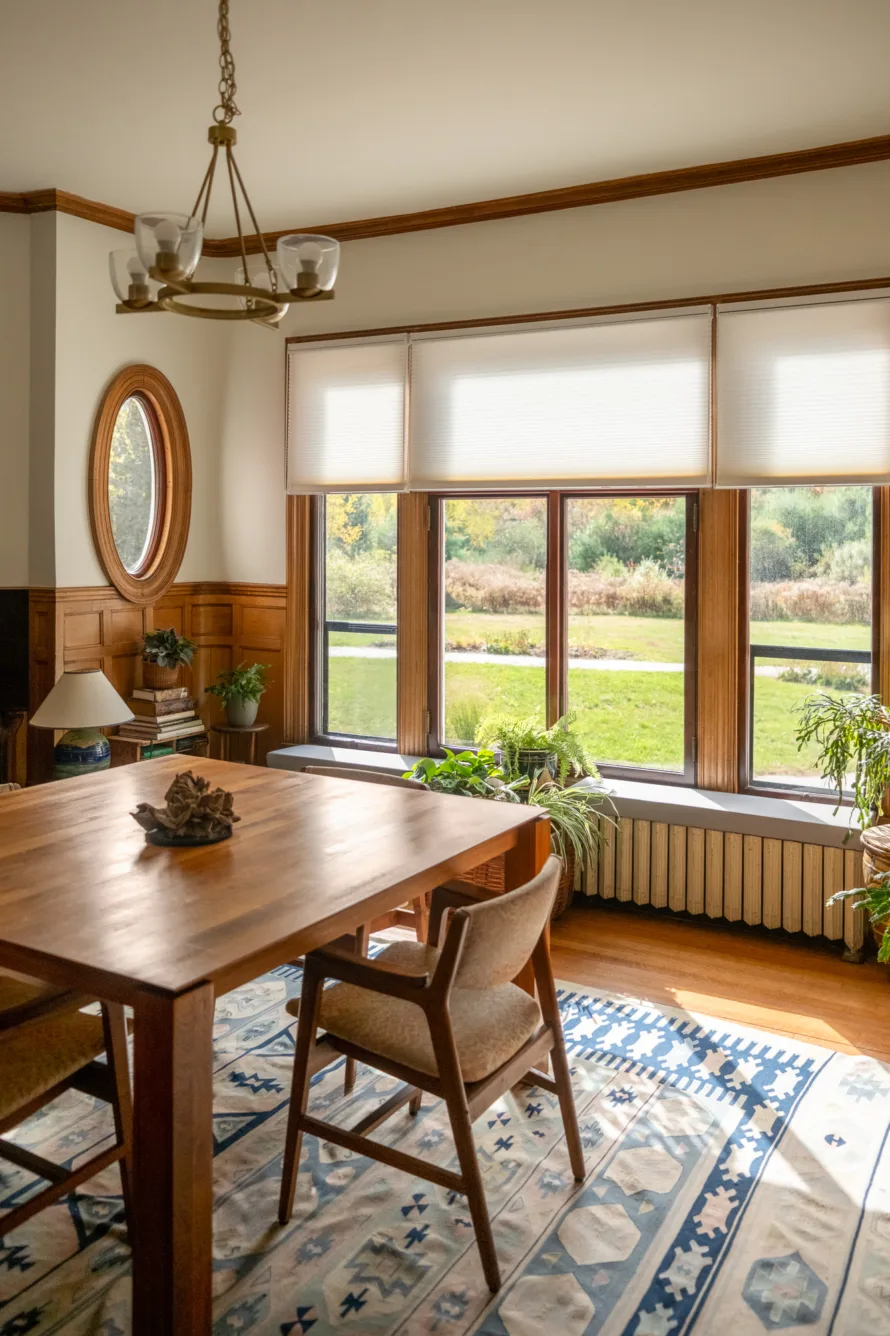 A bright dining room with large windows, wooden table and chairs, indoor plants, a blue-patterned rug, and a round mirror on the wall.