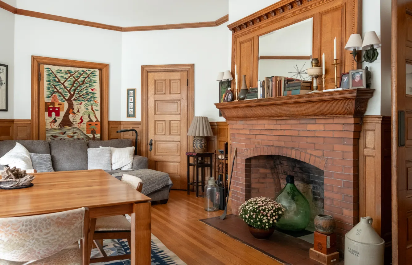 A cozy living room with a wooden dining table, gray sofa, brick fireplace with a mantle, books, candles, and plants. A colorful tapestry hangs on the wall by the window.