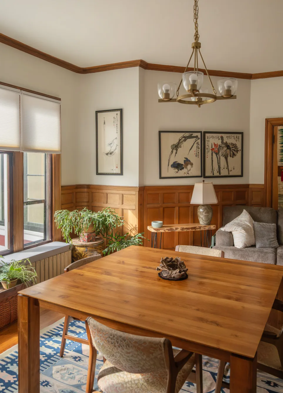 A cozy dining room with a wooden table, patterned rug, indoor plants by the window, and framed art on paneled walls, with a chandelier overhead and a lamp on a side table.
