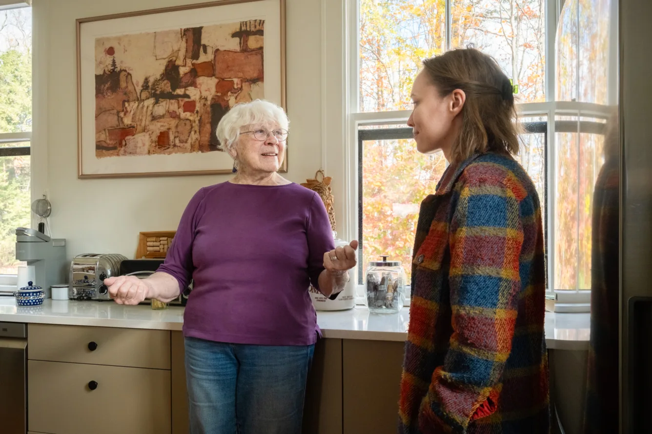 An older woman and a younger woman stand in a sunlit kitchen, engaged in conversation. The kitchen has large windows, modern cabinets, and a painting on the wall.