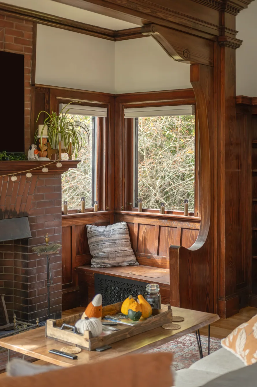Cozy living room with wood paneling, a brick fireplace, a window seat with pillows, and a coffee table decorated with gourds and autumn decor.