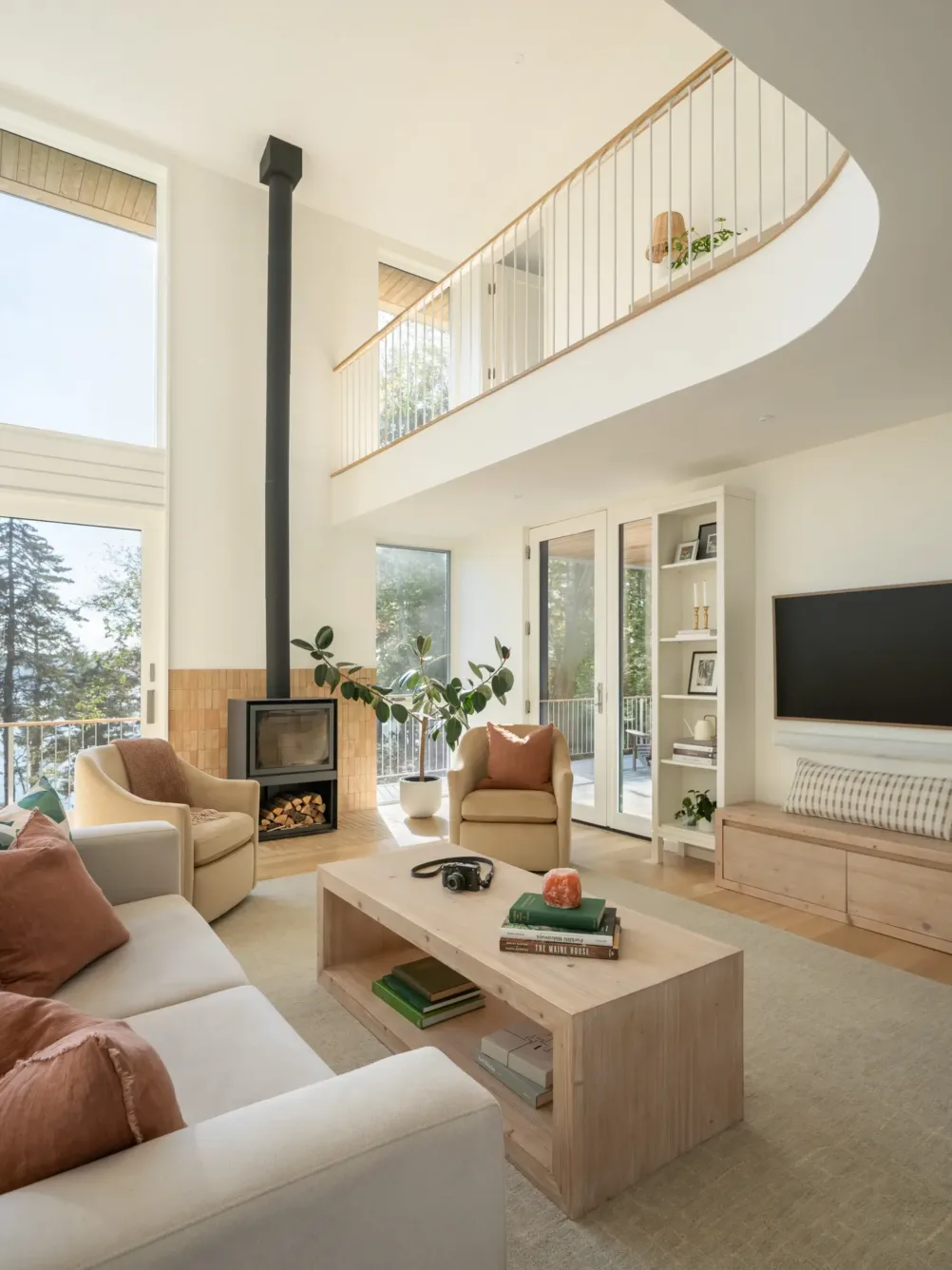 Modern, bright living room with large windows, light wood furniture, neutral sofas, orange cushions, a wood-burning stove, built-in shelves, and an open loft with railing above.