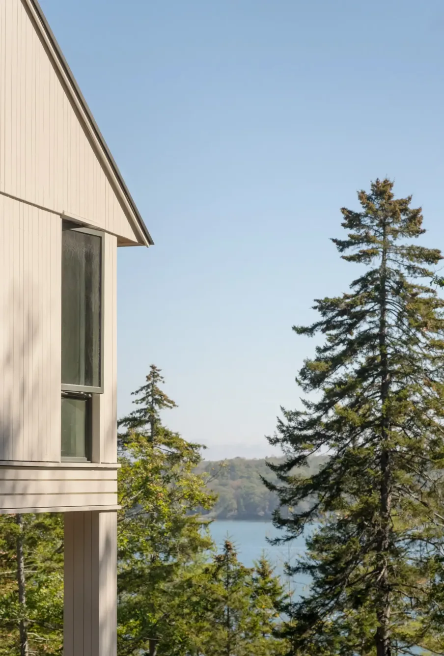A partial view of a modern house with large windows overlooks tall evergreen trees and a body of water under a clear blue sky.