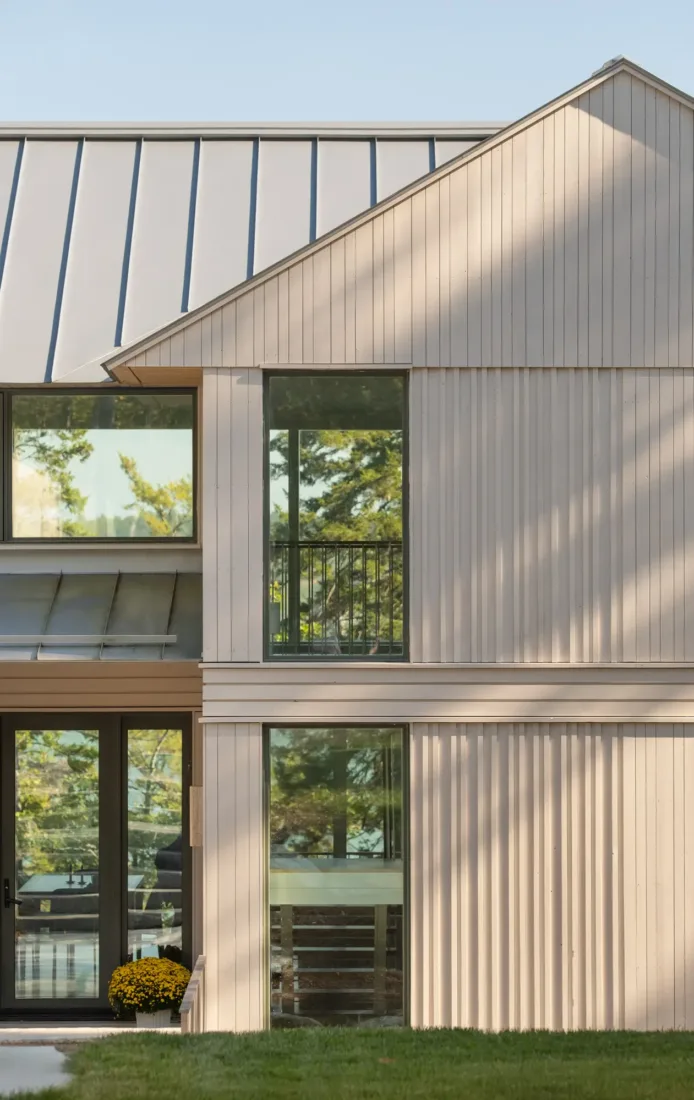Modern two-story house with light vertical wood siding, large windows reflecting trees, metal roof, and a small yellow potted plant by the glass door.
