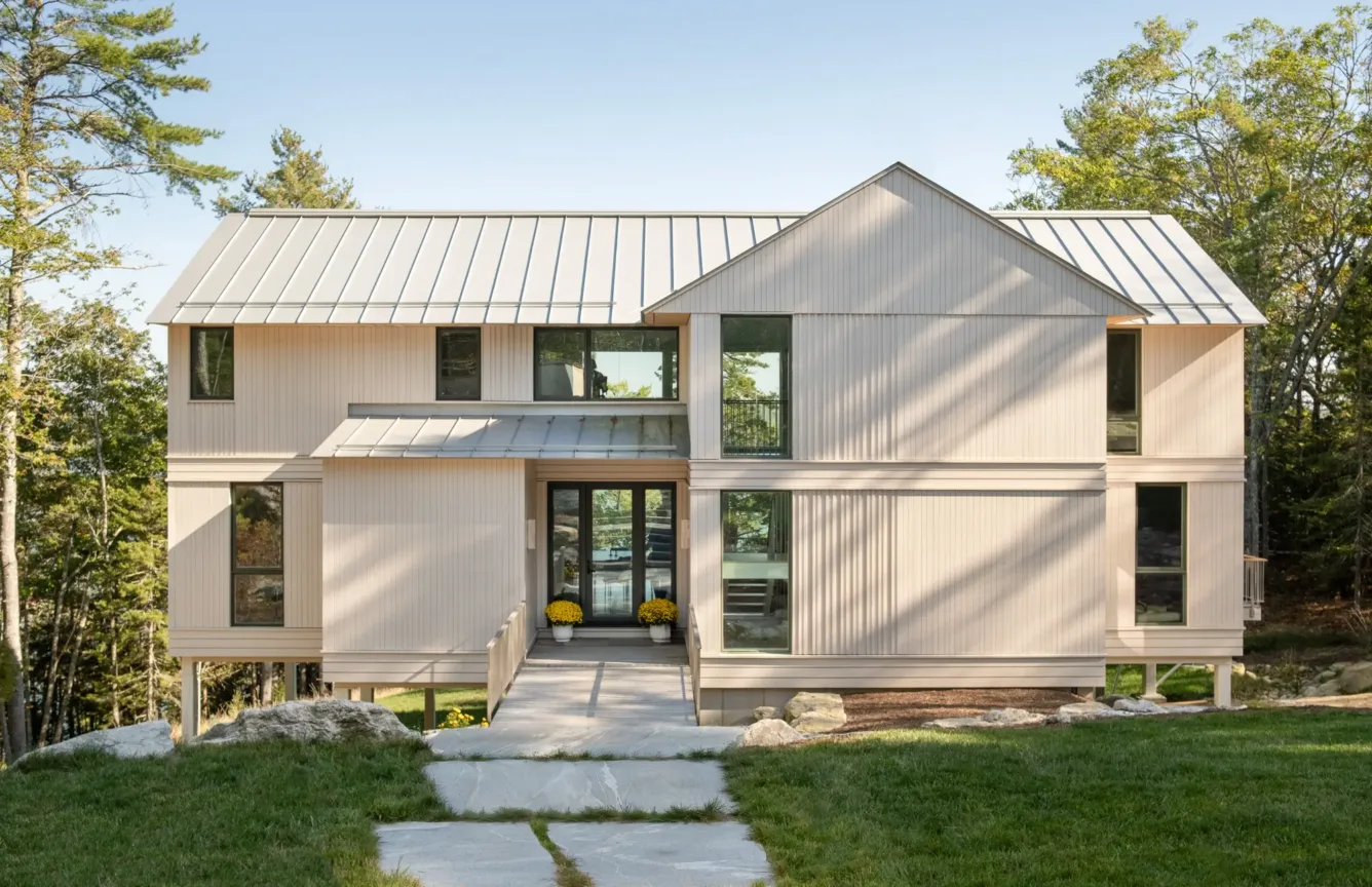 Modern two-story house with light wood siding, large windows, and metal roof; stone pathway leads to glass front doors with yellow potted flowers on the porch, surrounded by trees.