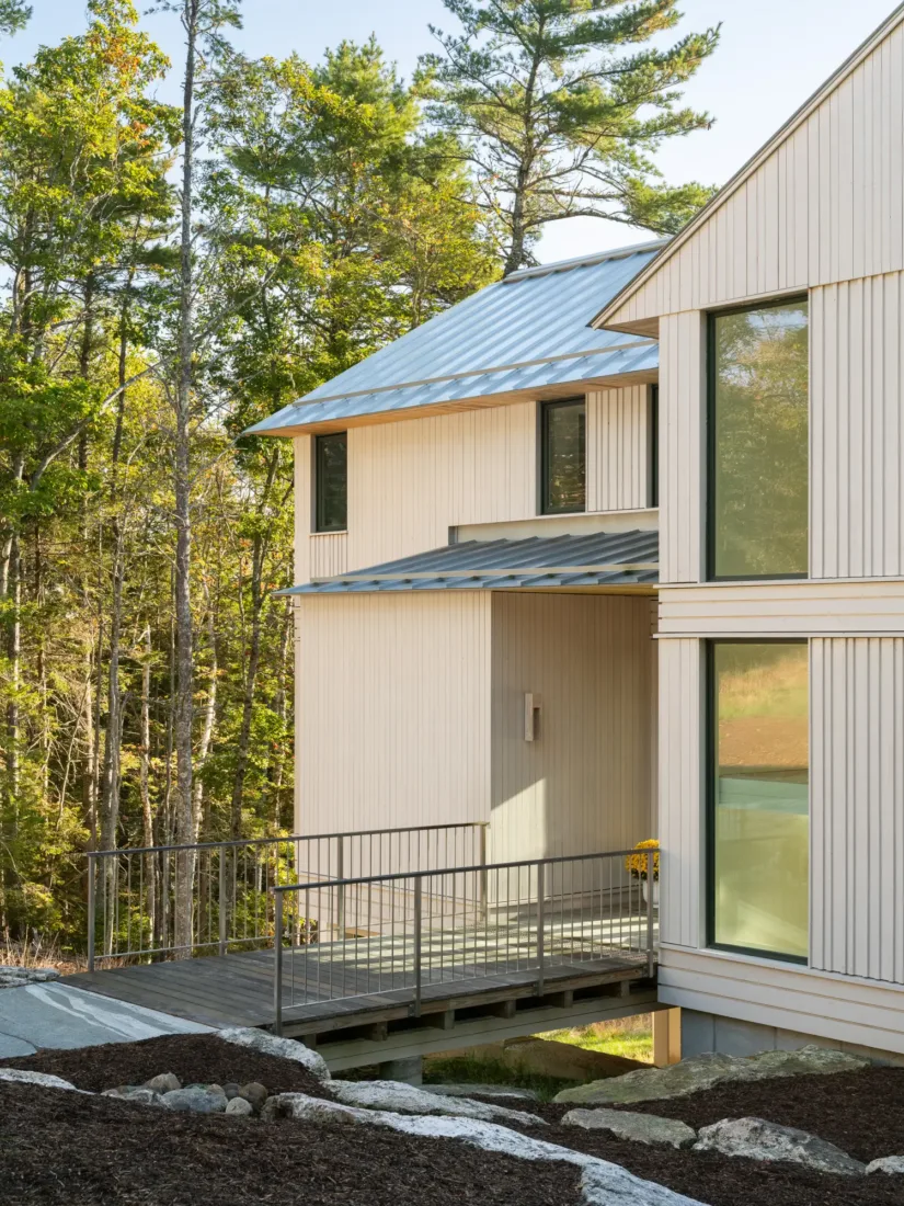 Modern house with light wood siding, large windows, a metal roof, and a small bridge-style walkway leading to the entrance, surrounded by trees and natural landscaping.