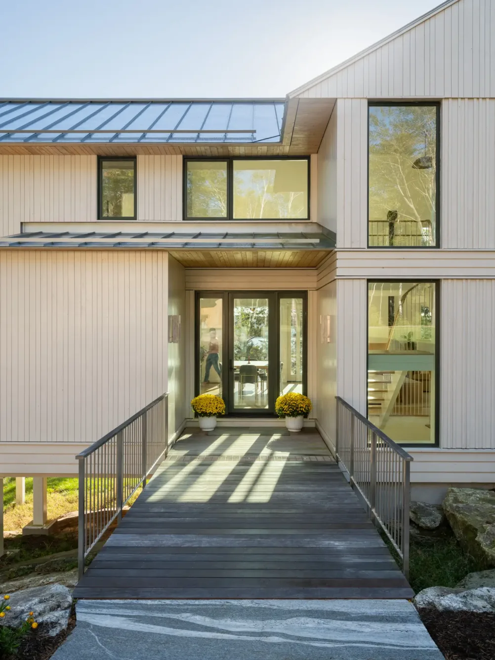 Modern house entrance with large glass doors, light wood siding, metal railings, and potted yellow flowers on either side of the walkway. Sunlight shines on the building.