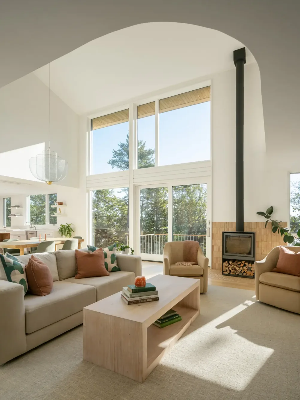 Bright living room with large windows, beige sofas with orange pillows, a light wooden coffee table, and a modern fireplace with stacked wood beside it. Sunlight floods the airy space.