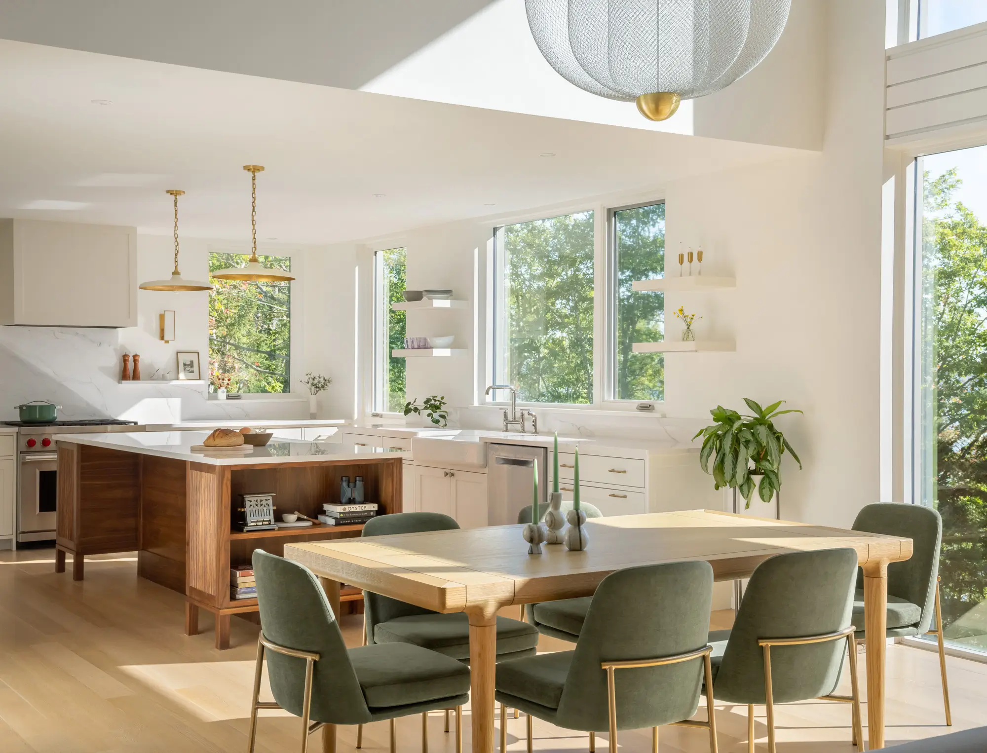 Bright, modern kitchen and dining area with large windows, green chairs, wooden table, white cabinets, plants, and gold accents, filled with natural light.