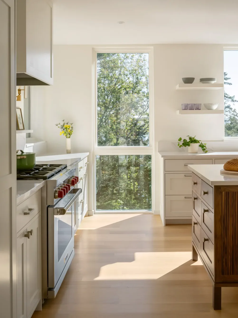 Bright, modern kitchen with white cabinets, wooden island, and large window letting in sunlight and views of green trees outside. Minimalist decor with a few plants and dishes on shelves.