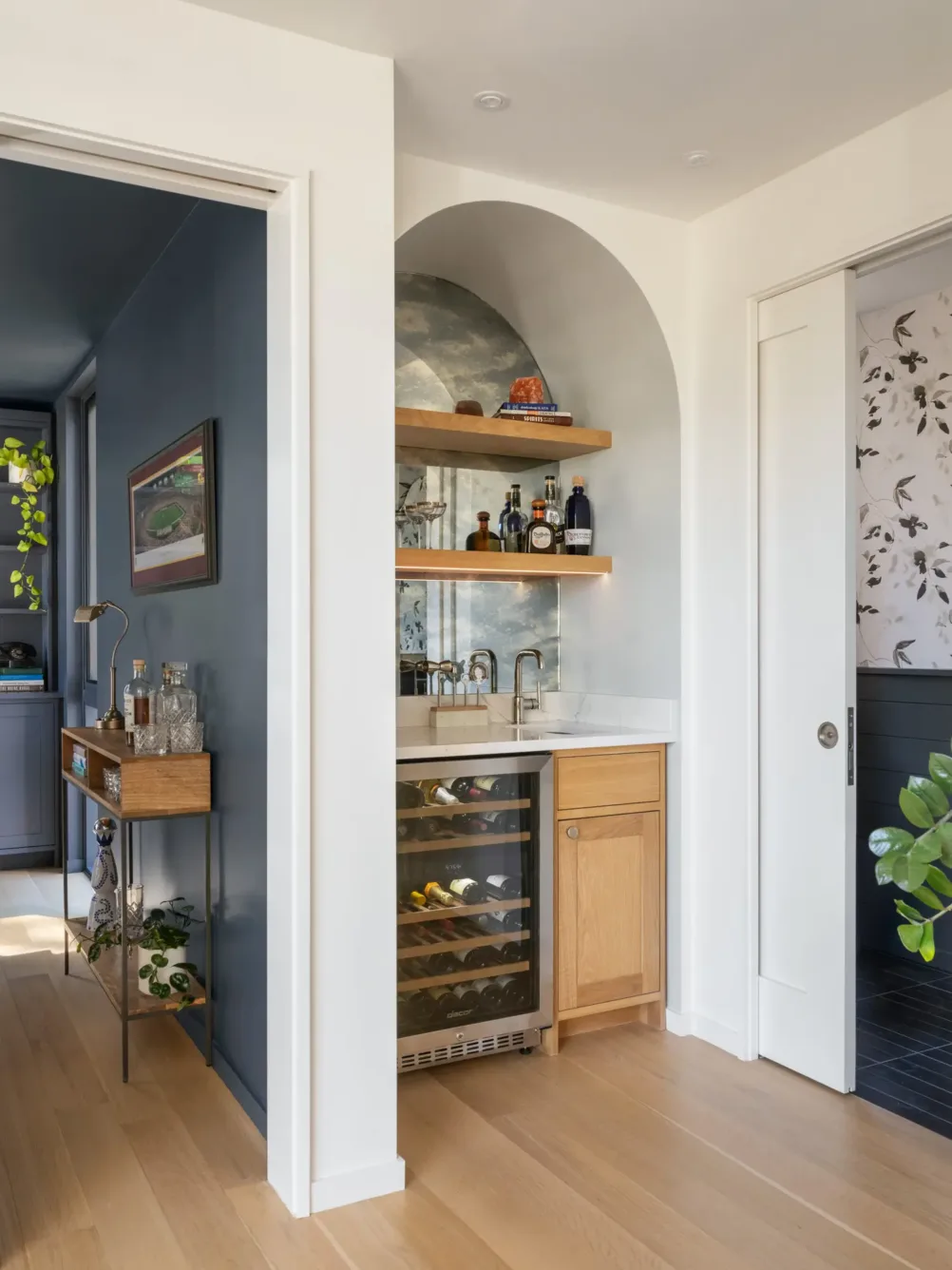A small home bar with a mini fridge, wood cabinets, open shelves with bottles, a sink, and an arched niche with a cloud mural, set between two rooms with wood floors.