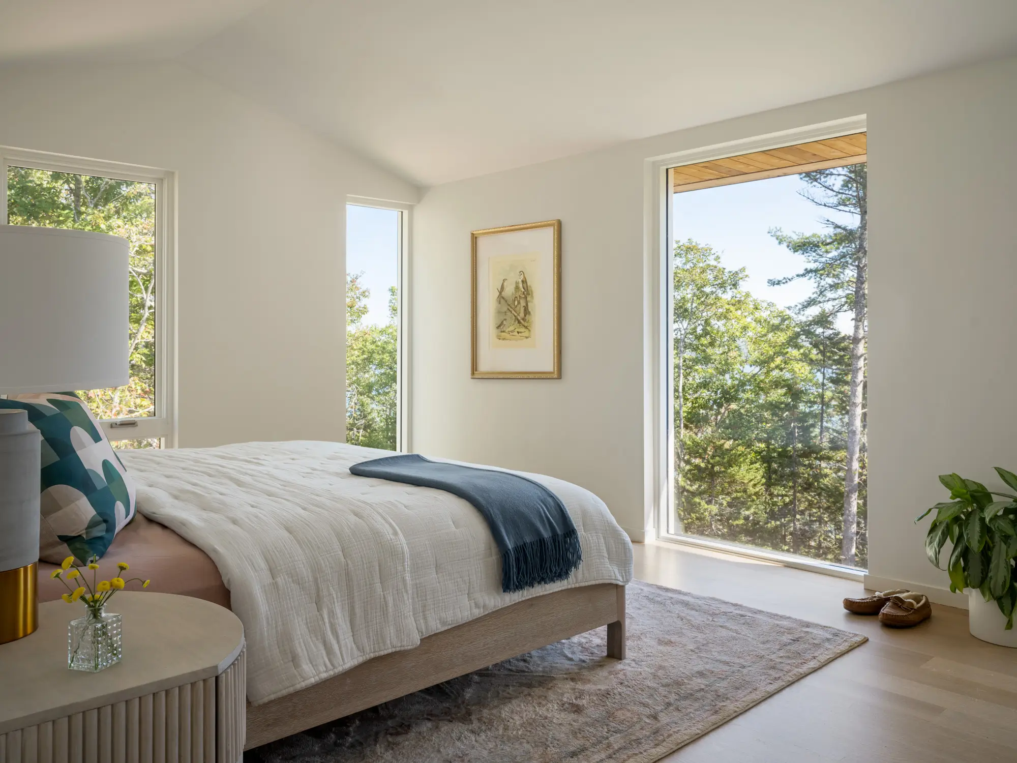 A bright, modern bedroom with large windows, a cozy bed with white bedding and a blue throw, a nightstand, a plant, and trees visible outside.